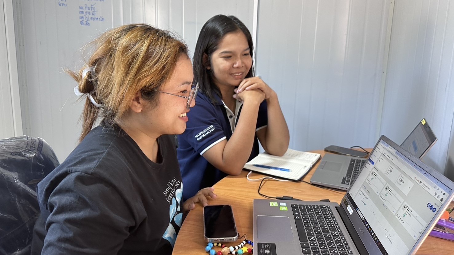 Two woman smile as they look at a laptop computer on a brown table. 